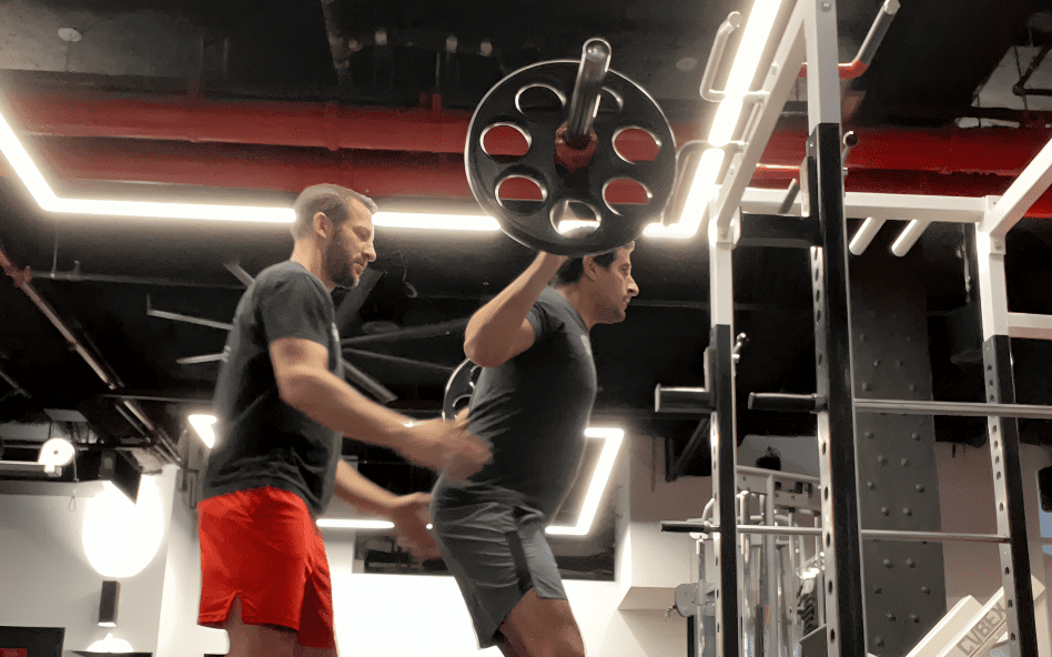 Man squatting with barbell, trainer assisting.
