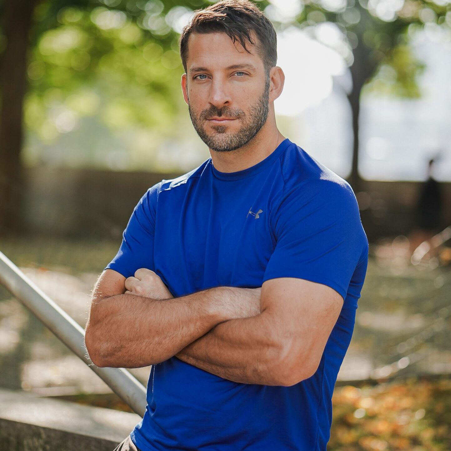 Fit man in blue shirt posing outdoors with arms crossed.